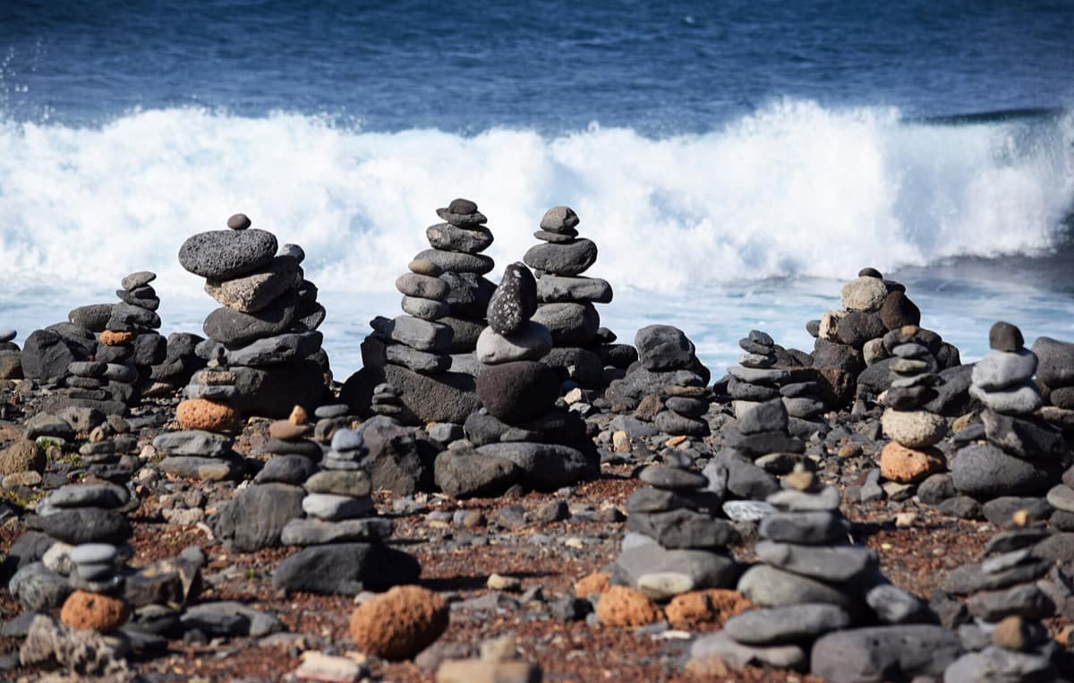 Des empilements de galets sur une plage volcanique sombre, devant une vague qui déferle.